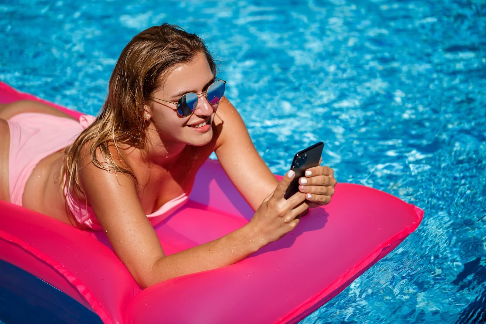 Sexy woman with a phone in a swimsuit lies on a pink inflatable mattress in the pool. Relax by the pool on a hot summer sunny day. Vacation concept