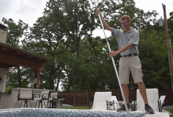 Emerald Pool Care technician skimming a backyard pool under large trees during fall pool maintenance in Fort Worth, TX