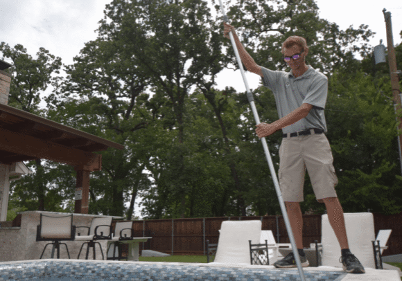 Emerald Pool Care technician skimming a backyard pool under large trees during fall pool maintenance in Fort Worth, TX